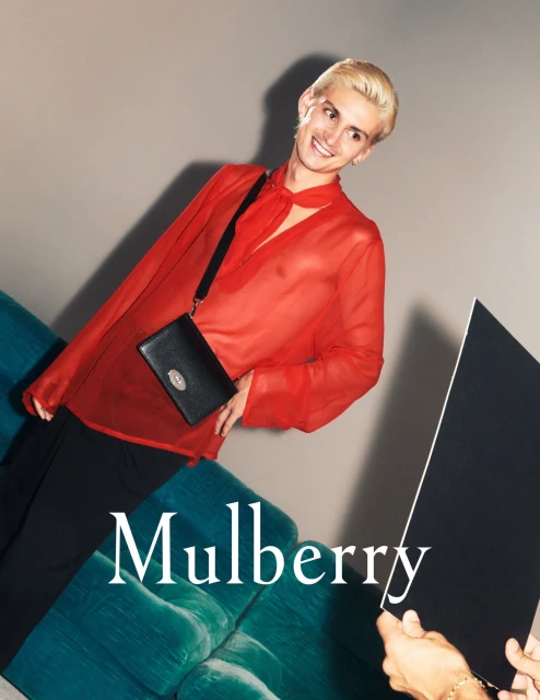 A fashionable person with short blonde hair, wearing a red shirt and black pants and holding a black bag, poses indoors.