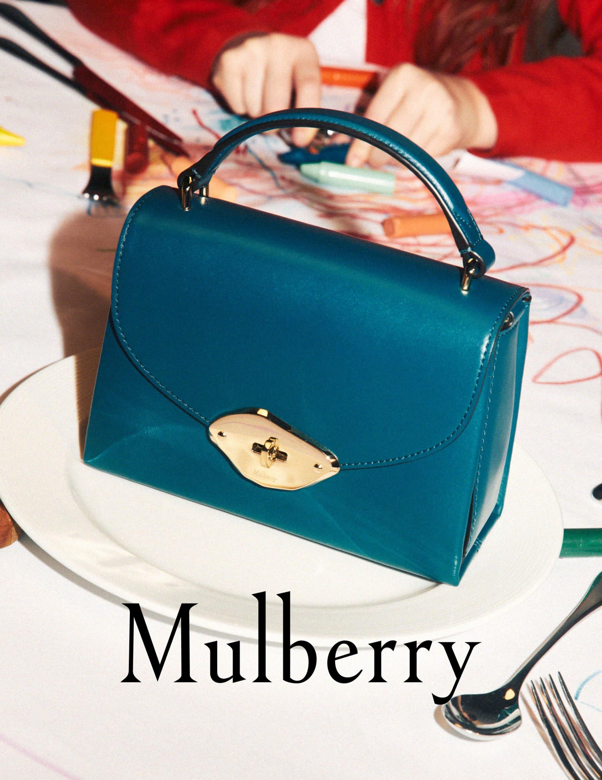Turquoise Mulberry handbag on a white plate, with a child drawing in the background, showcasing fashion and family lifestyle.