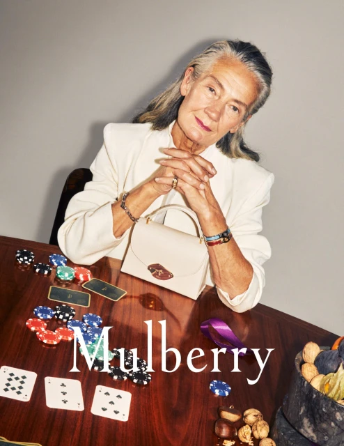 Elegant older woman poses at a poker table with a cream handbag, chips, and cards. The Mulberry brand name is visible in the frame.