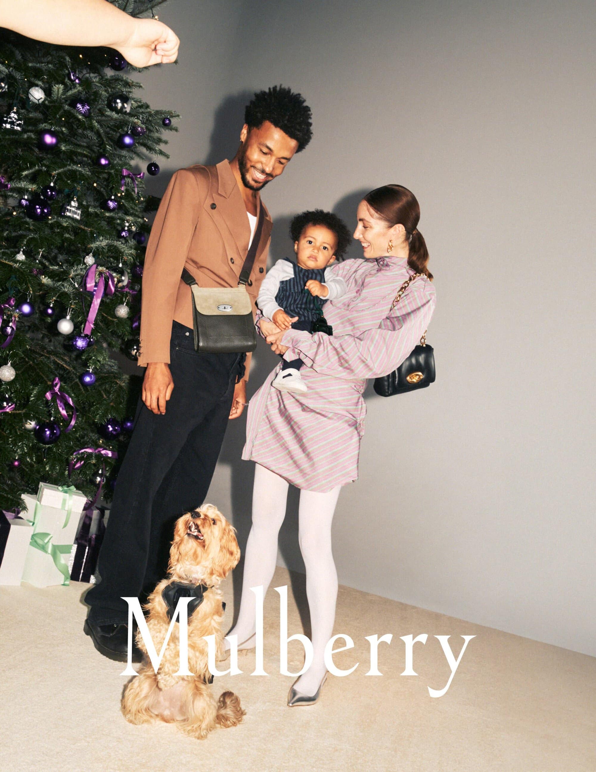 Family of three with a dog in front of a Christmas tree, posing with gifts. The family is stylishly dressed in warm tones.