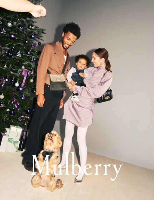 Family of three with a dog in front of a Christmas tree, posing with gifts. The family is stylishly dressed in warm tones.