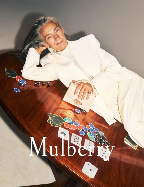 Mature woman in a white suit lays on a table with playing cards, dice, and poker chips near a white purse on a neutral background.