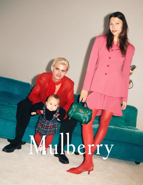 Fashion shot of an adult woman in a pink suit, an adult man with a child and a green purse, posing indoors on a turquoise sofa.