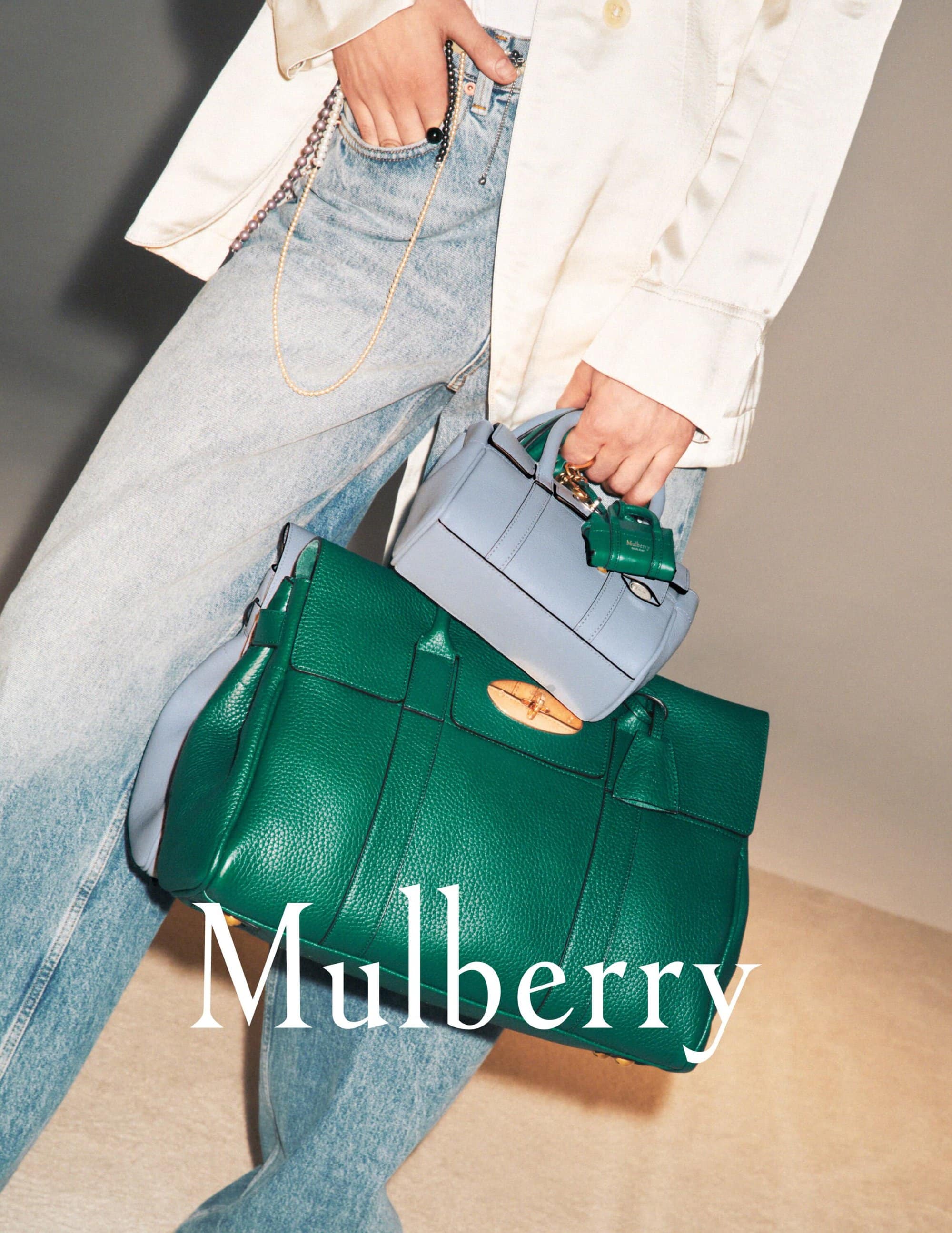 Close-up studio shot of a green leather Mulberry handbag and a light blue leather bag with an adult hand and light blue denim jeans.
