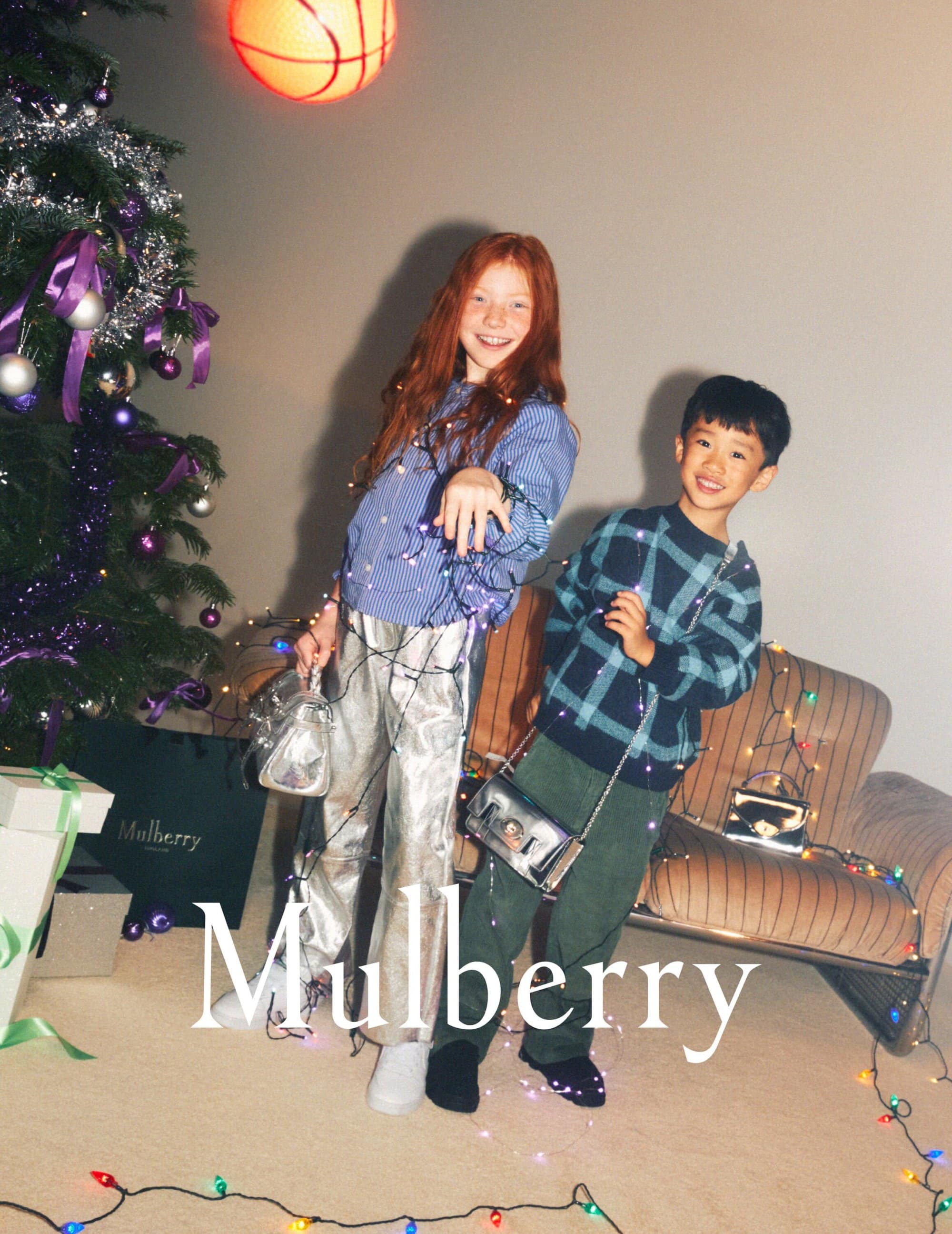 Two smiling children, a girl with red hair and a boy with dark hair, pose with handbags and Christmas lights in a festive indoor setting.