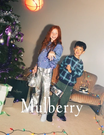 Two smiling children, a girl with red hair and a boy with dark hair, pose with handbags and Christmas lights in a festive indoor setting.