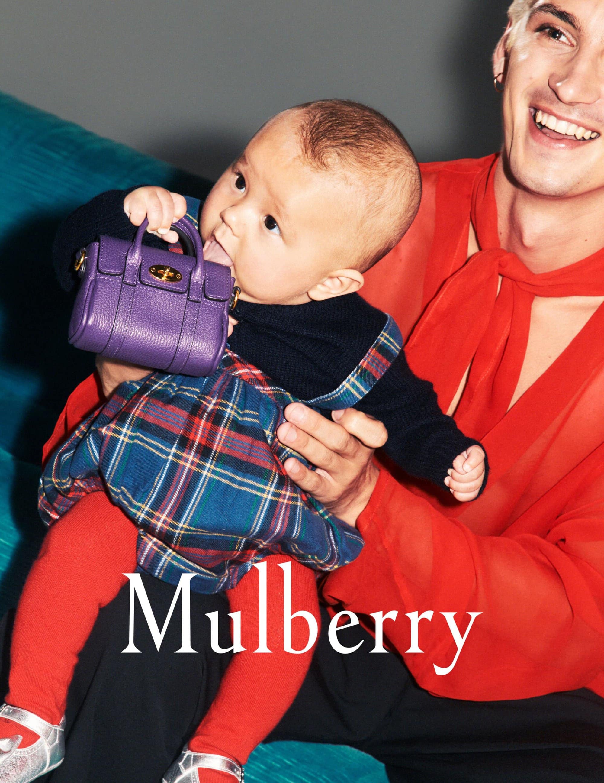A baby in a plaid skirt and red tights sitting in an adult’s lap, chewing on a purple Mulberry handbag. The adult wears a red shirt.