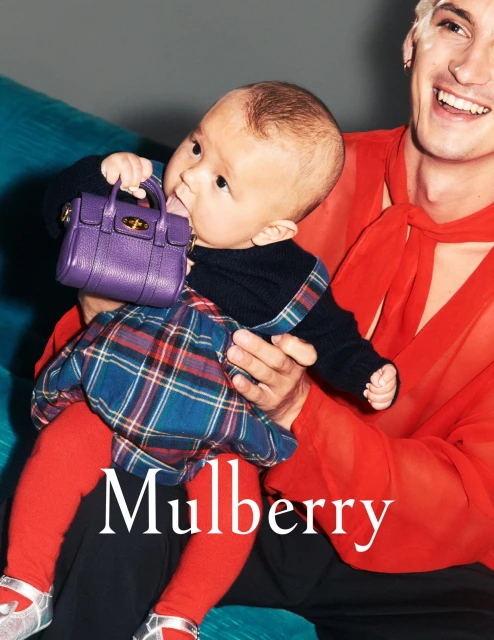 A baby in a plaid skirt and red tights sitting in an adult’s lap, chewing on a purple Mulberry handbag. The adult wears a red shirt.