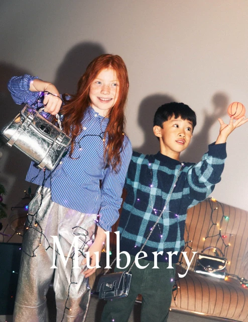 Two young children, a girl with red hair and a boy with dark hair, pose with fashion bags against a neutral background with string lights.