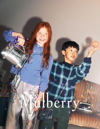 Two young children, a girl with red hair and a boy with dark hair, pose with fashion bags against a neutral background with string lights.