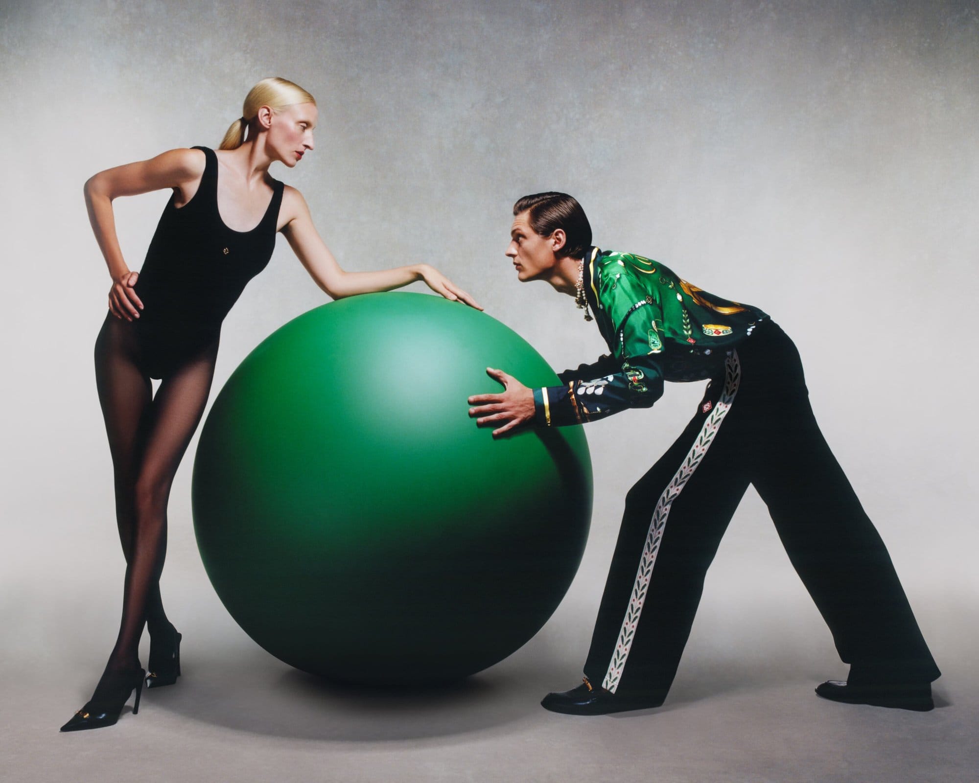 Two fashion models pose in studio with a large green ball. One woman wears a black leotard, tights, and heels; a man wears a green and gold jacket.