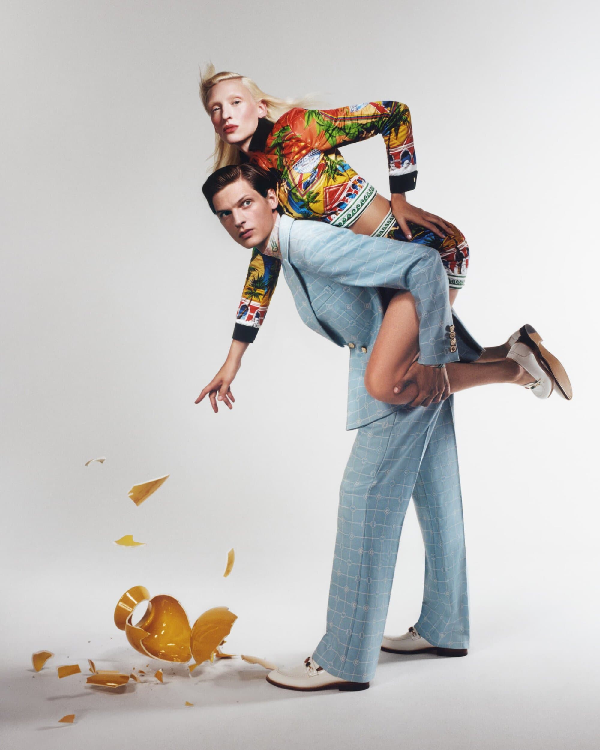Fashion shot of a man in a suit holding up a woman wearing a crop top and skirt, next to a broken vase in a studio setting.