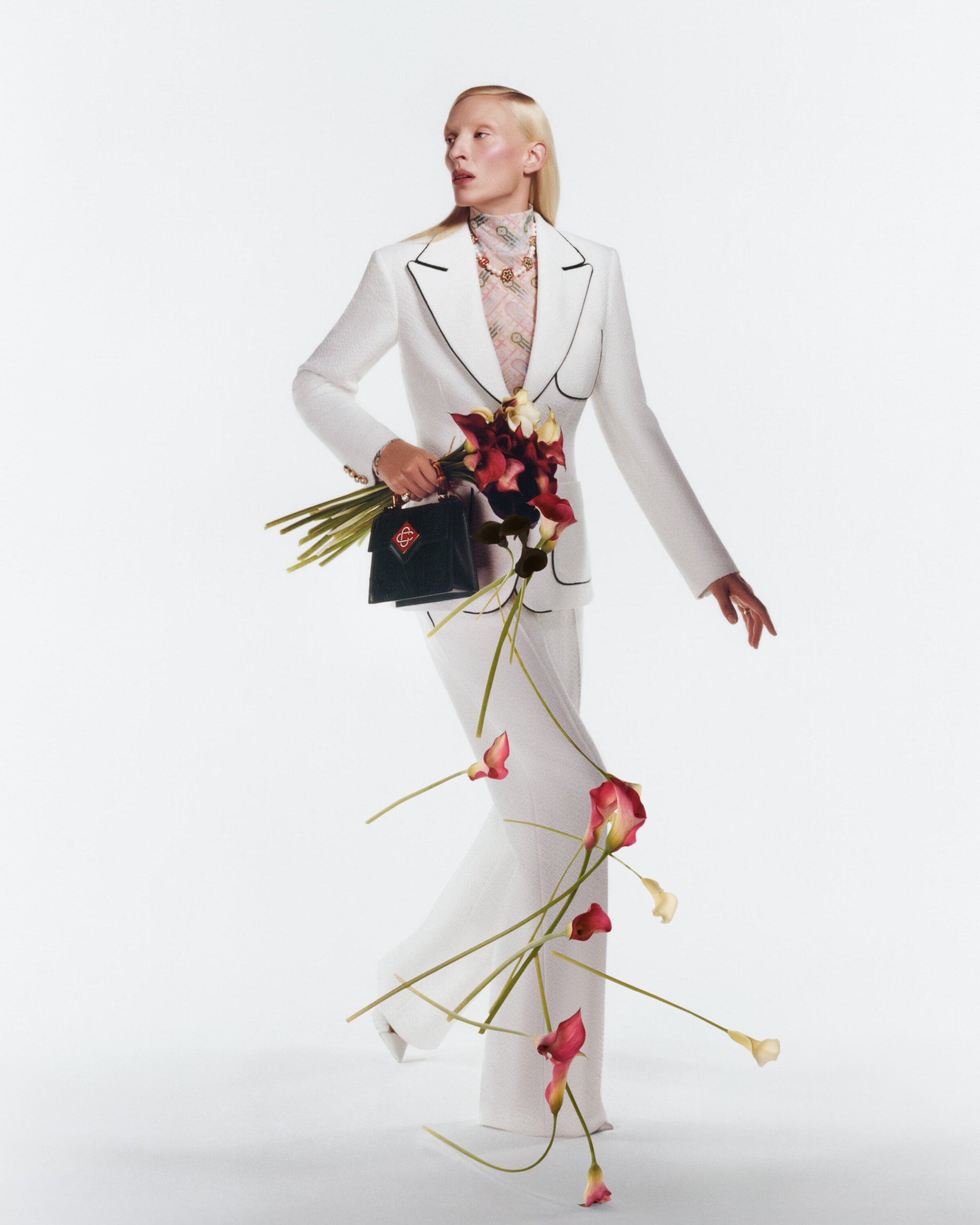 Fashion shot of a fair-skinned model in a white suit with black trim, holding a dark bag with lilies cascading down, against a white backdrop.