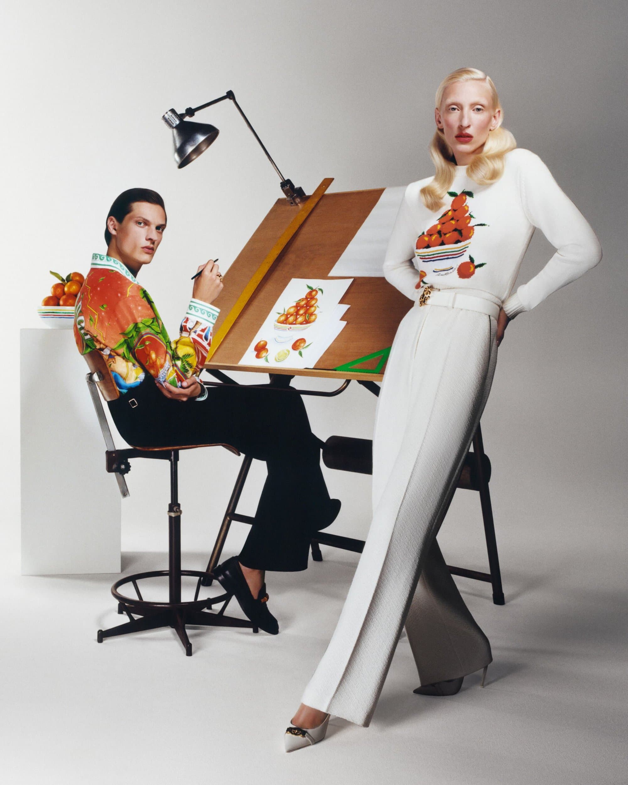 Fashion editorial shot of two fashion models posing indoors with fruit-themed outfits, one sketching at a desk, bright lighting, white backdrop.