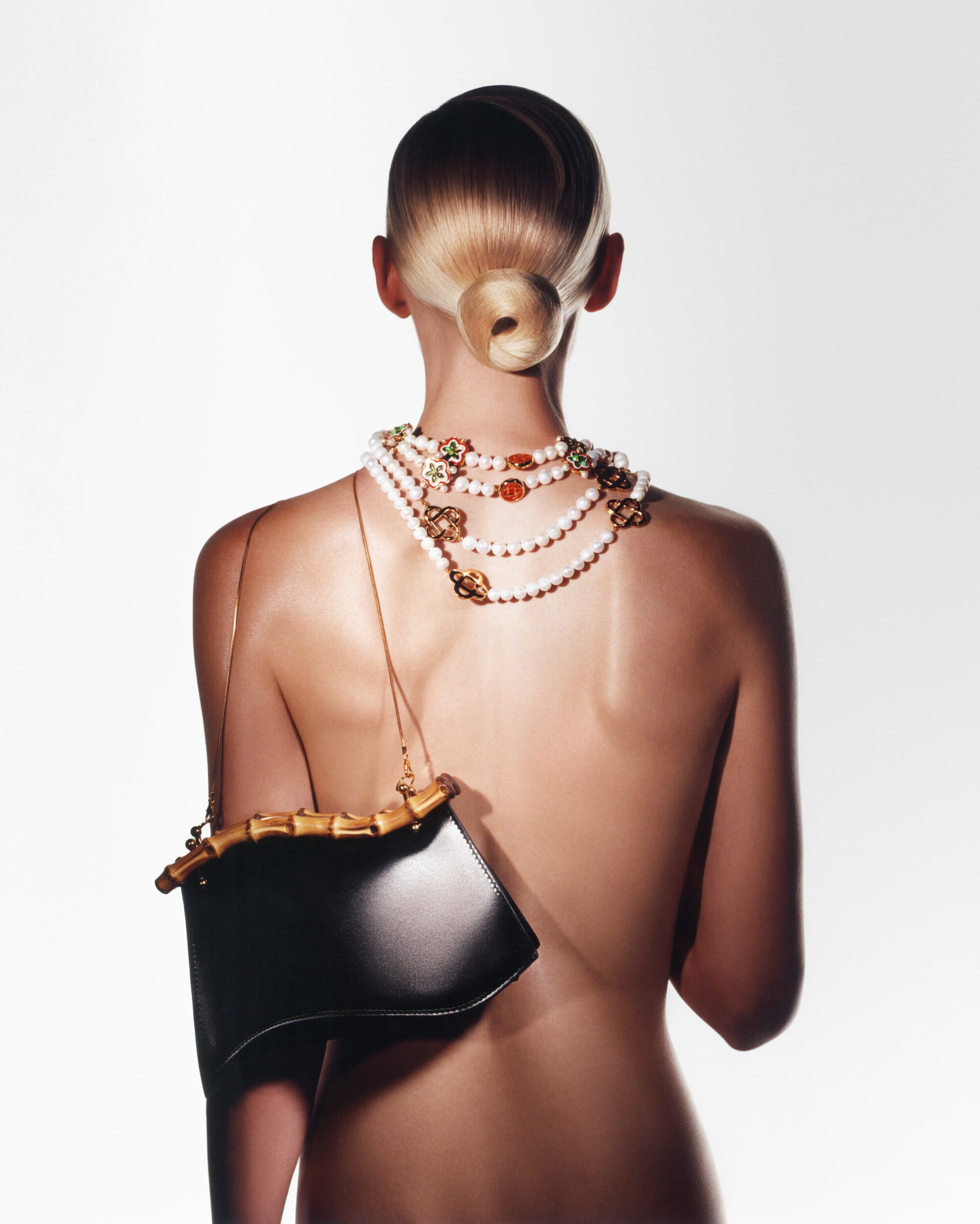 Back view of a fair-skinned, topless person with an elegant updo, adorned with layered pearl and gold necklaces, and a black bamboo-handled purse.