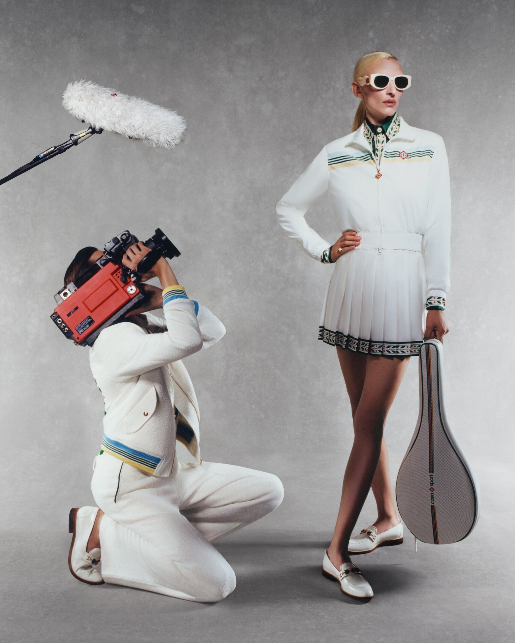 Stylish woman in a white tennis outfit poses with a videographer using vintage camera equipment in a studio against a grey backdrop.