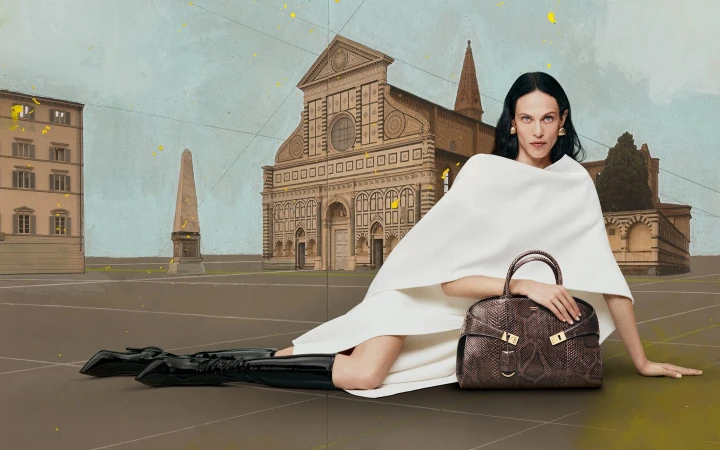 Fashion shot of an adult female in a white garment seated and posing with a handbag. The backdrop is a painted landscape.