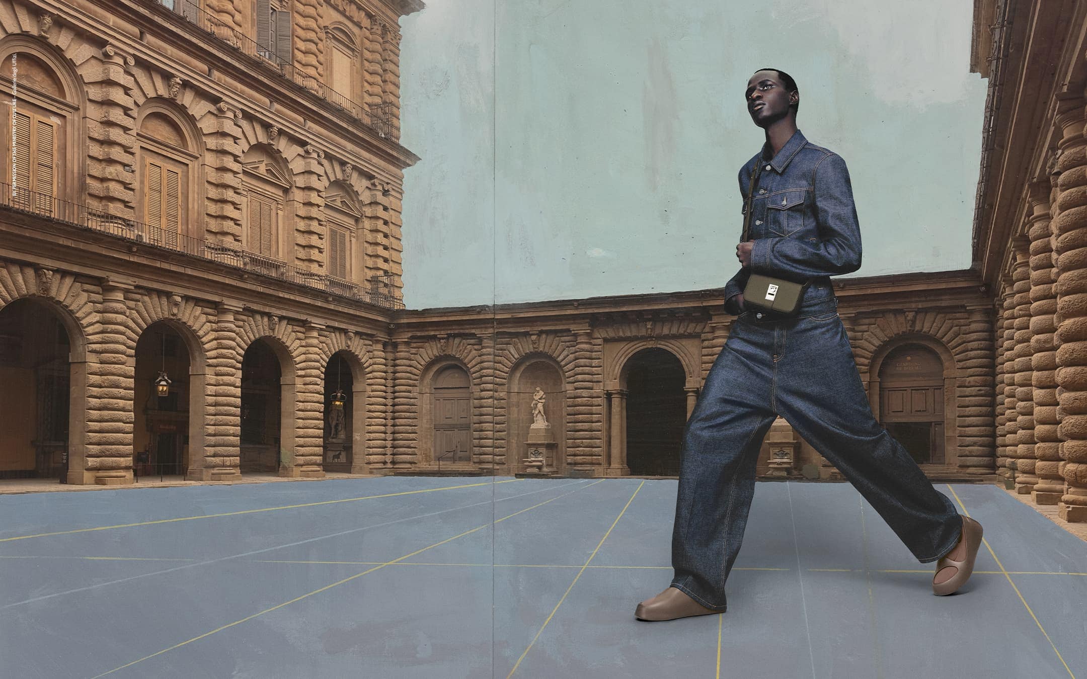 Adult male strides across blue courtyard wearing denim jacket, baggy jeans, shoulder bag, and neutral loafers, against palazzo backdrop.
