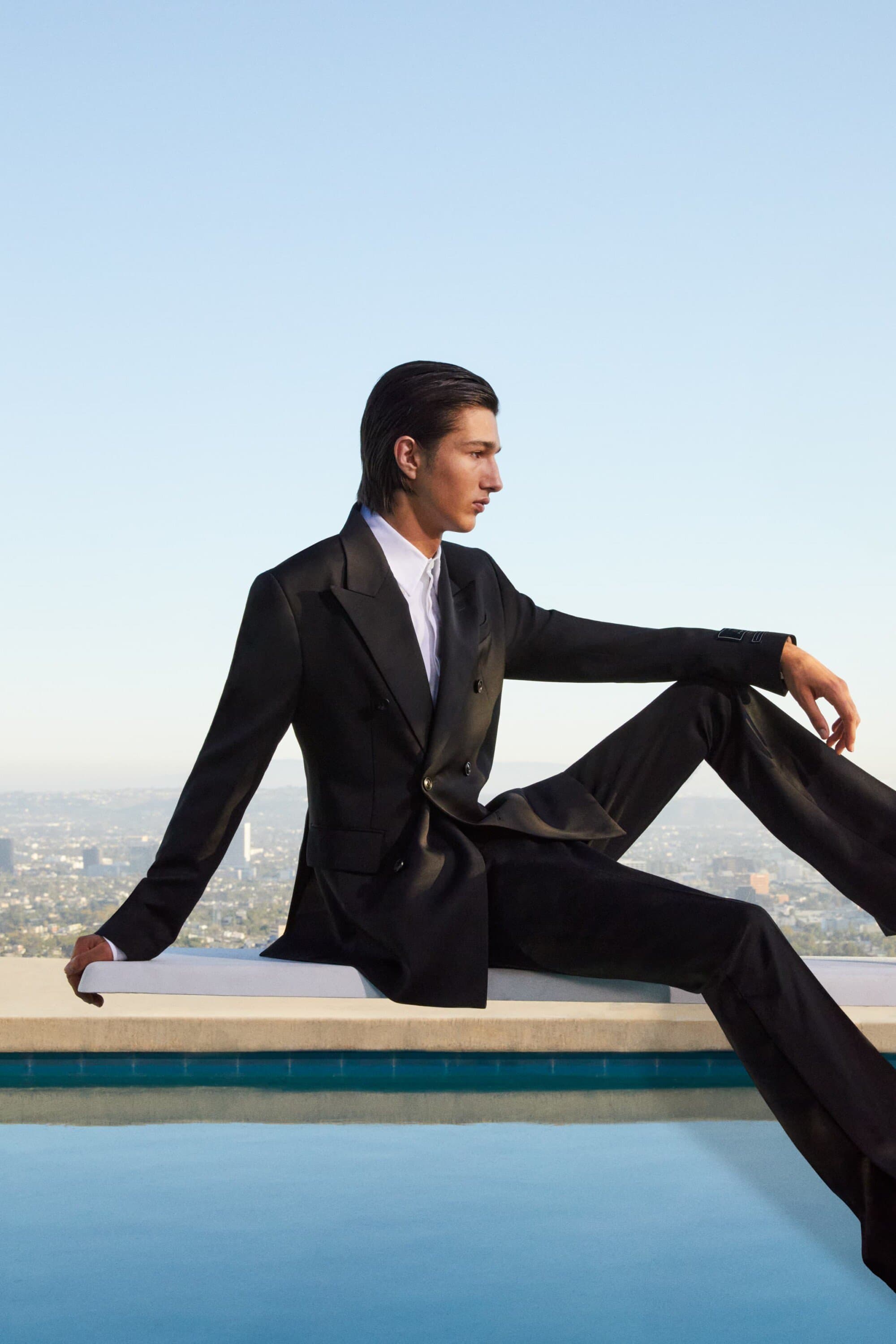 A fashionable young adult male model poses in a black double-breasted suit near a pool, with an urban skyline in the background.
