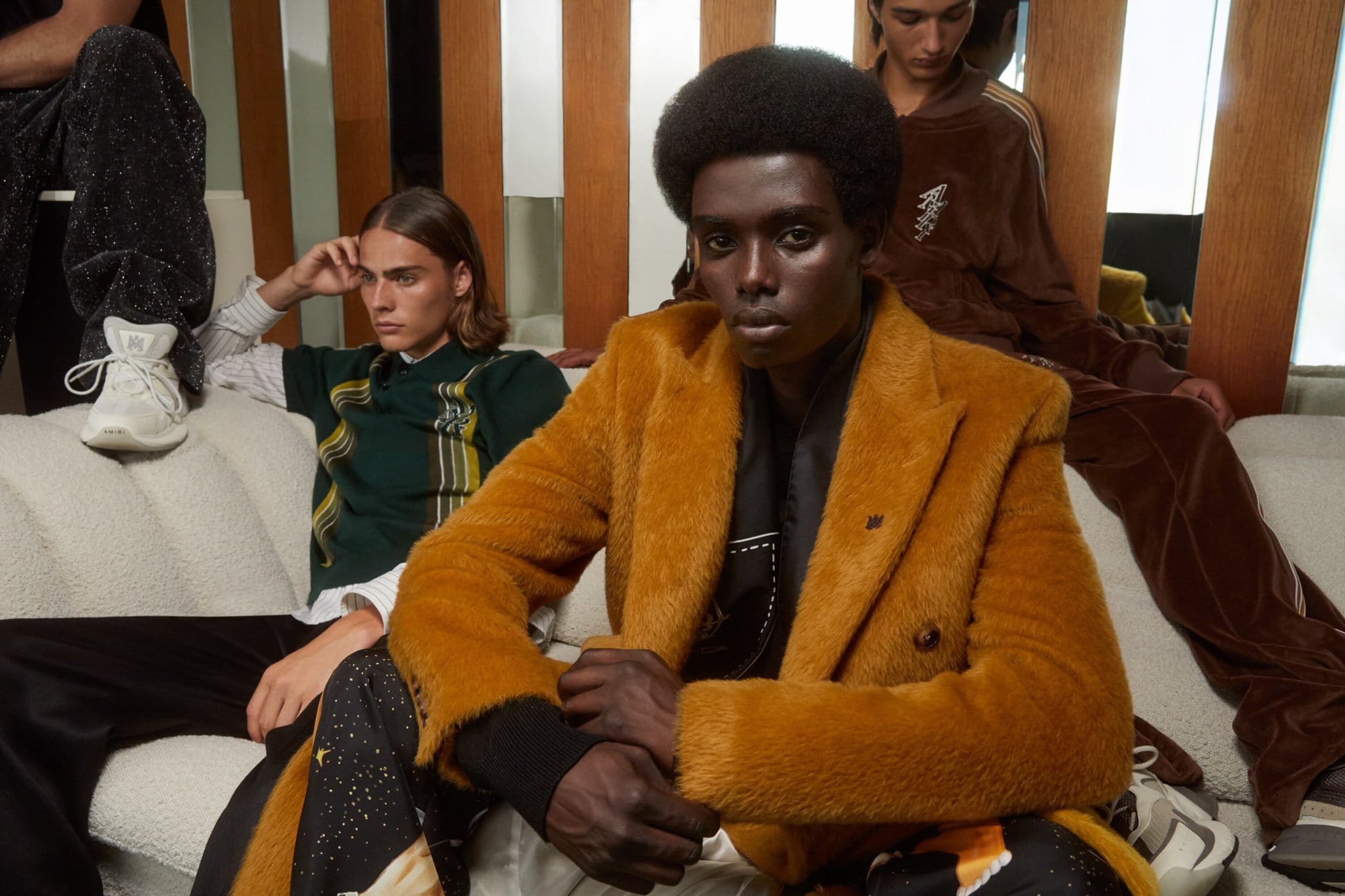 A Black model with an afro wearing a textured mustard yellow overcoat sits on a cream sofa with two other models in an Amiri ad campaign.