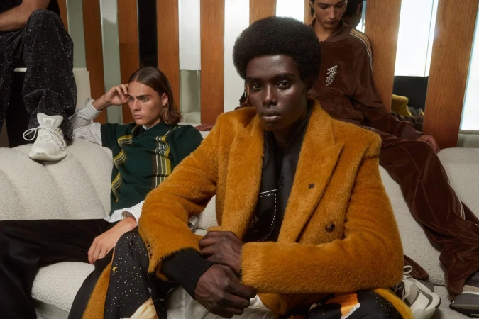 A Black model with an afro wearing a textured mustard yellow overcoat sits on a cream sofa with two other models in an Amiri ad campaign.