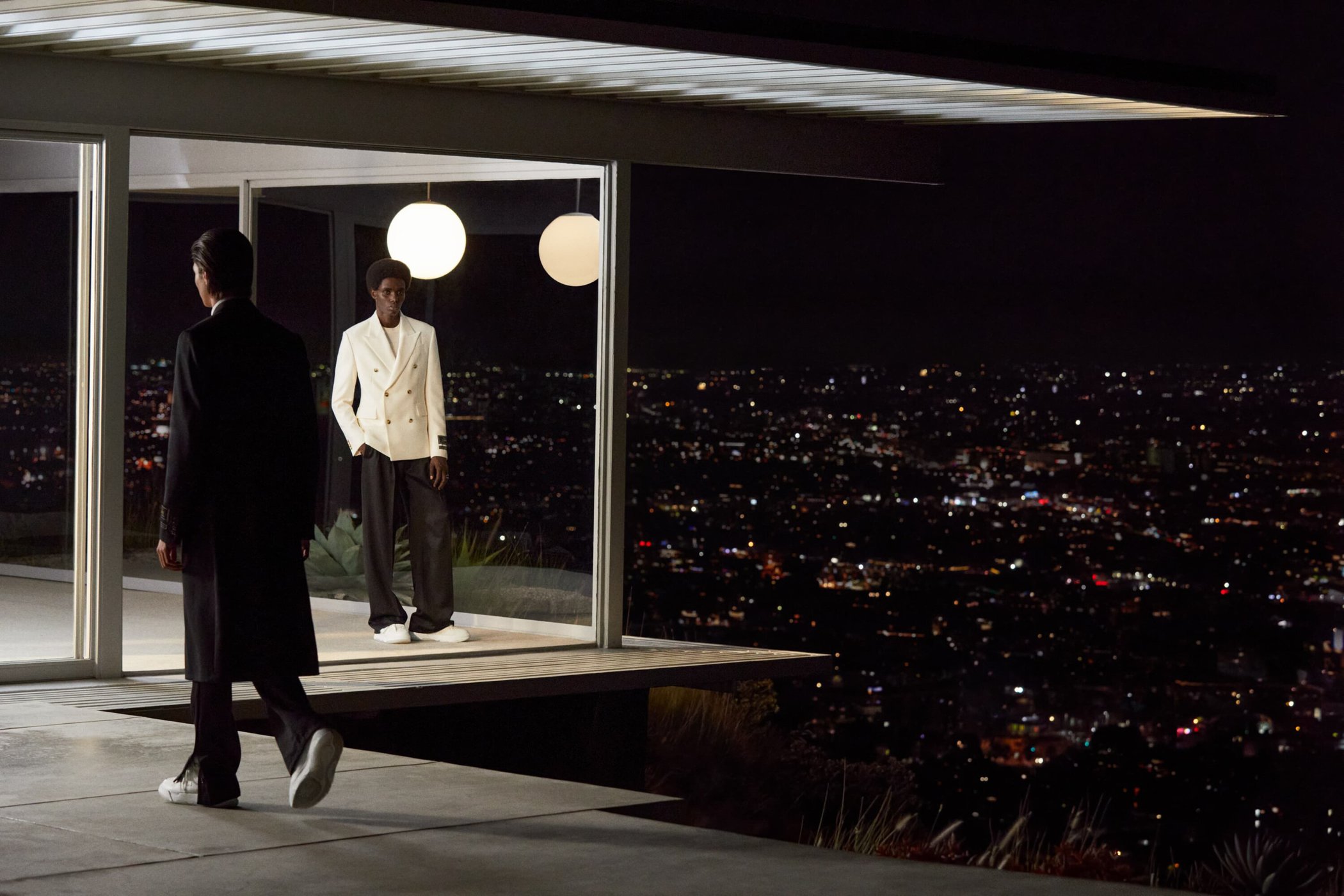 Two stylishly dressed men pose in front of a modern home at night overlooking a lit cityscape, showcasing fashion and architecture.