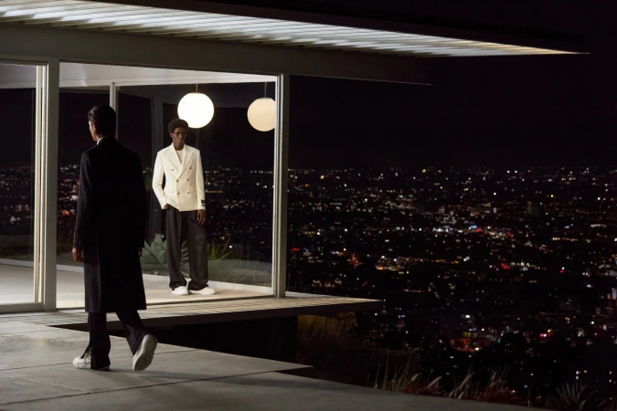 Two stylishly dressed men pose in front of a modern home at night overlooking a lit cityscape, showcasing fashion and architecture.