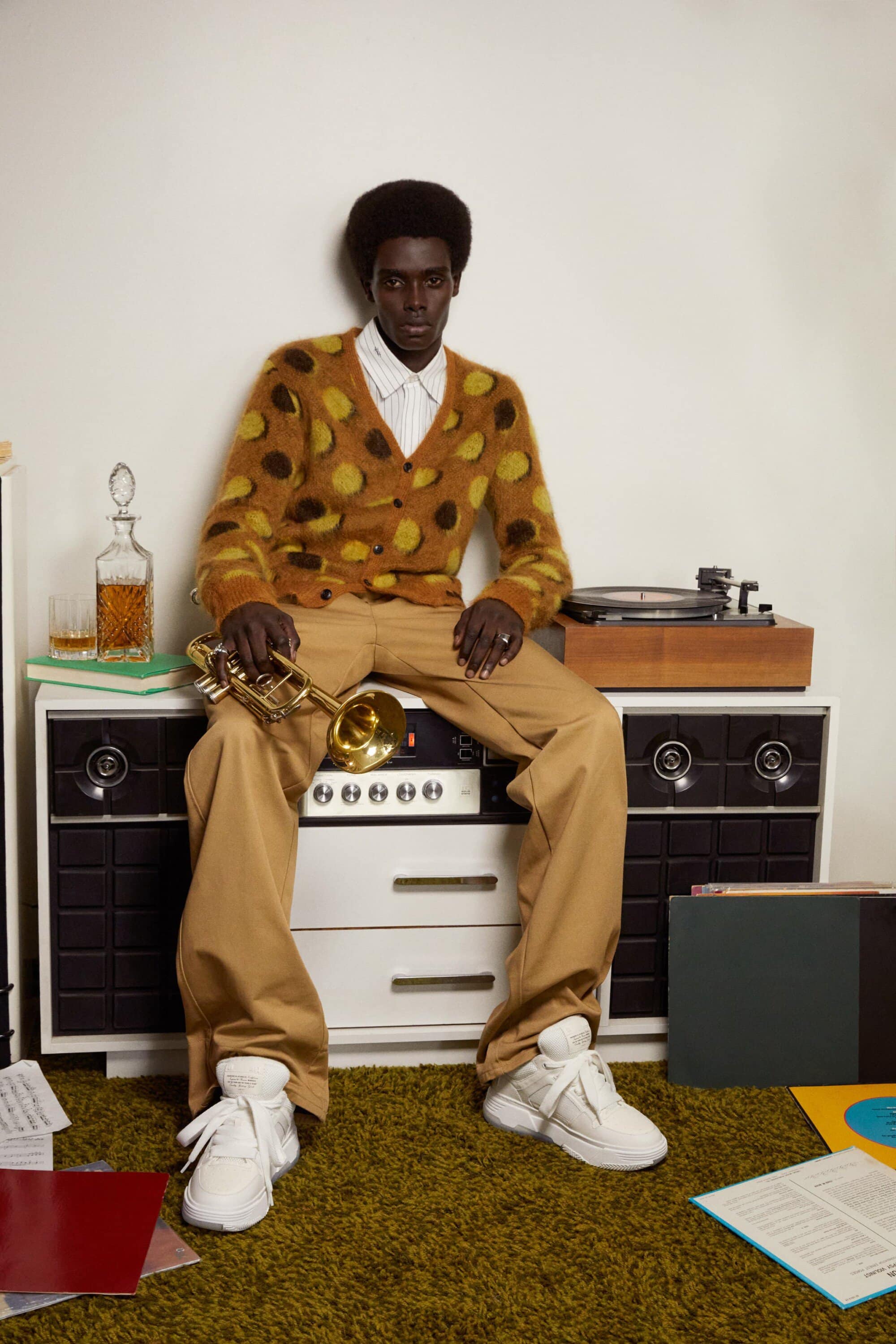Man in patterned cardigan sits on vintage audio equipment holding trumpet indoors, wearing khaki pants and white sneakers on green carpet.