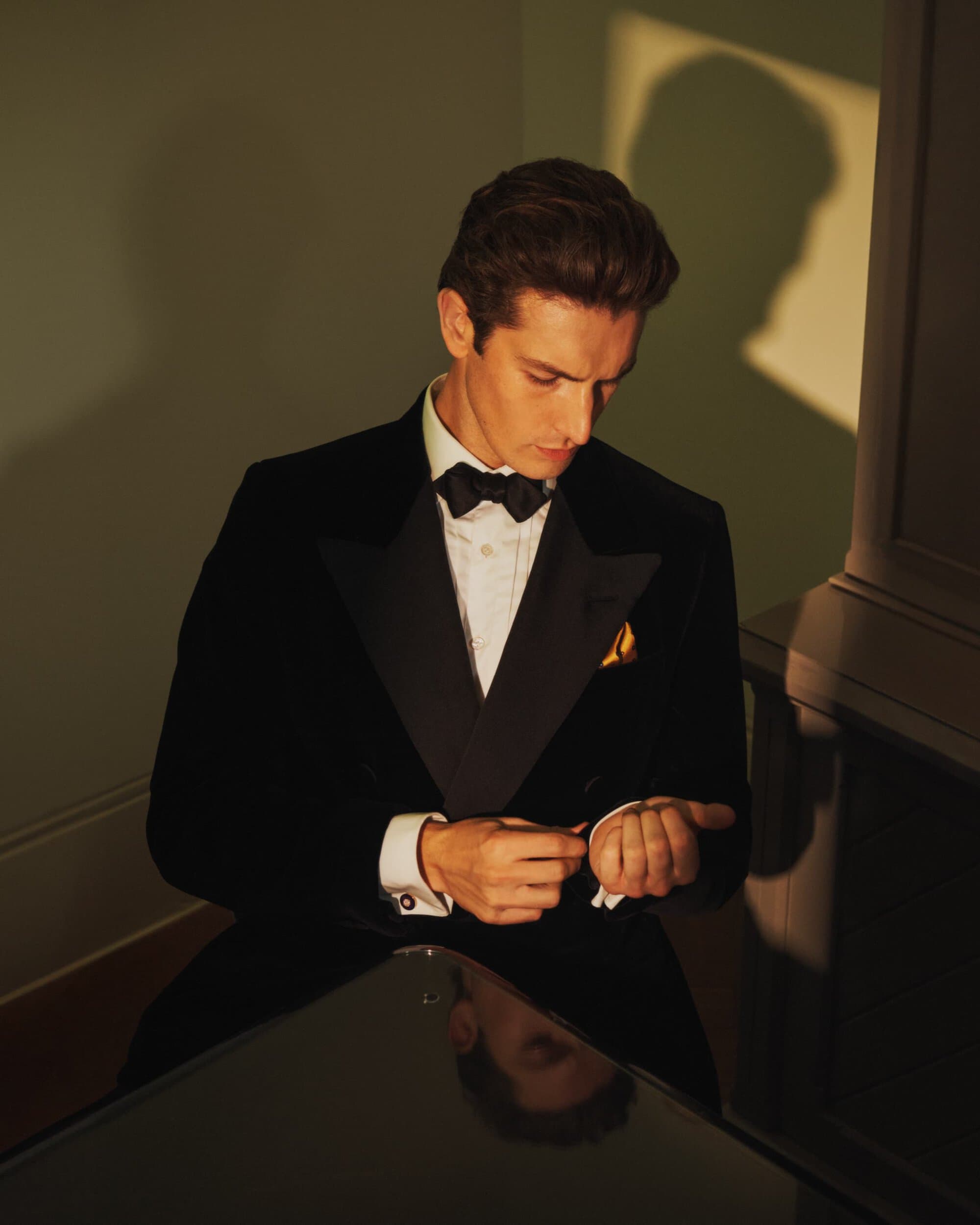 Portrait of a handsome man in a black tuxedo and bow tie adjusting his cufflink in an indoor setting with neutral lighting.