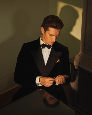 Portrait of a handsome man in a black tuxedo and bow tie adjusting his cufflink in an indoor setting with neutral lighting.
