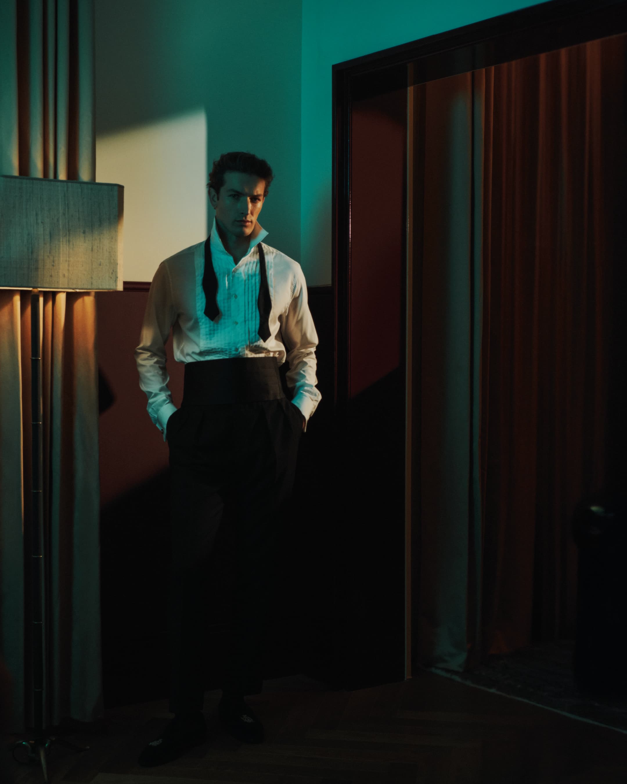 Portrait of a young adult male wearing a white shirt and black dress pants, standing in an indoor setting with a lamp and door.