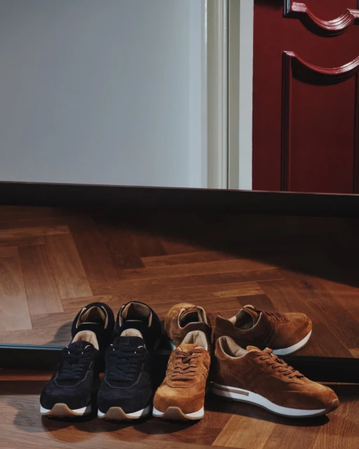 Two pairs of unworn brown suede leather sneakers and one pair of black sneakers, arranged on a parquet wood floor near a doorway.