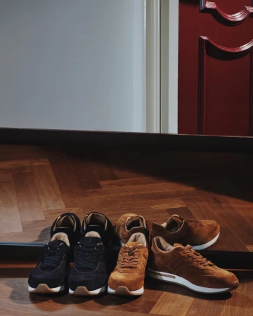 Two pairs of unworn brown suede leather sneakers and one pair of black sneakers, arranged on a parquet wood floor near a doorway.