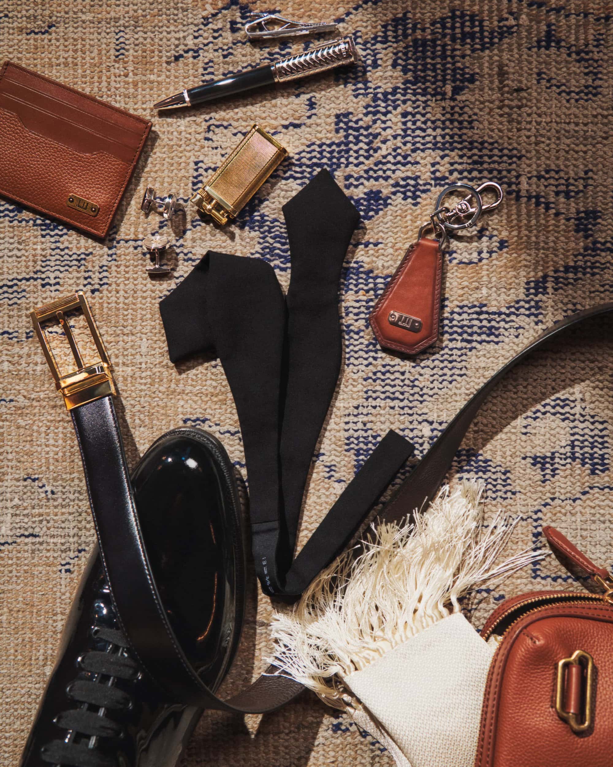 Flat lay of men's accessories including a wallet, pen, lighter, bow tie, shoes, belt, and bag, arranged on a patterned rug.