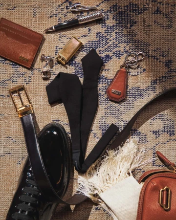 Flat lay of men's accessories including a wallet, pen, lighter, bow tie, shoes, belt, and bag, arranged on a patterned rug.