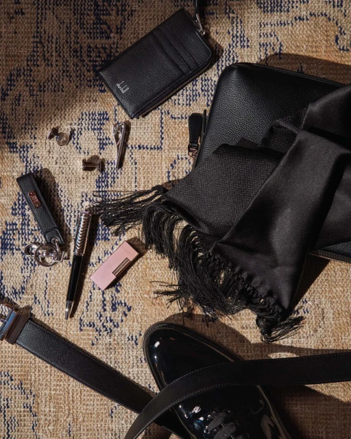 Still life of men's accessories on patterned rug: leather wallet, shoes, belt, pen, scarf, keychains, cufflinks, and lighter.