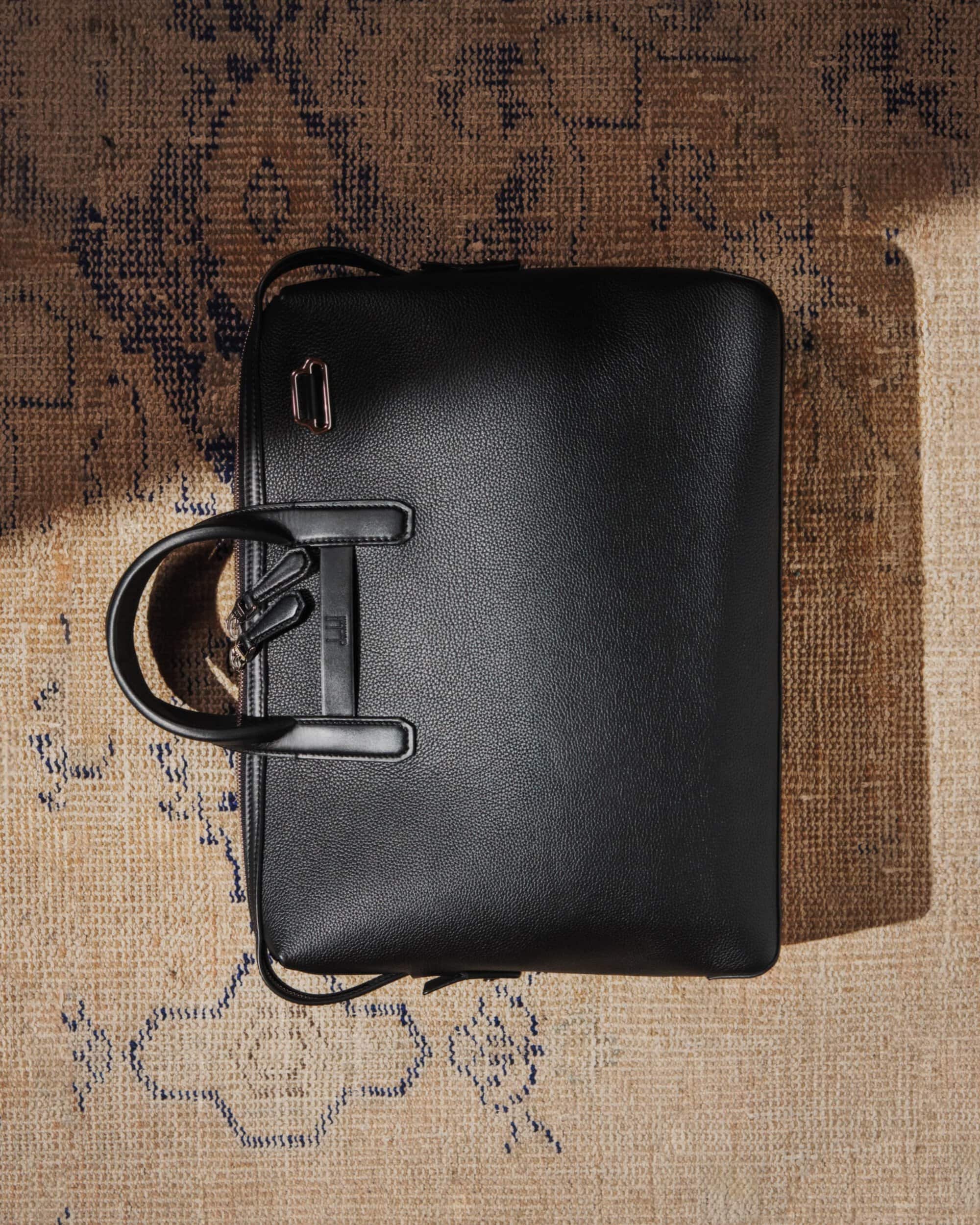 Overhead studio shot of a black leather laptop bag with handles, on an ornate rug with neutral tones and blue accents.