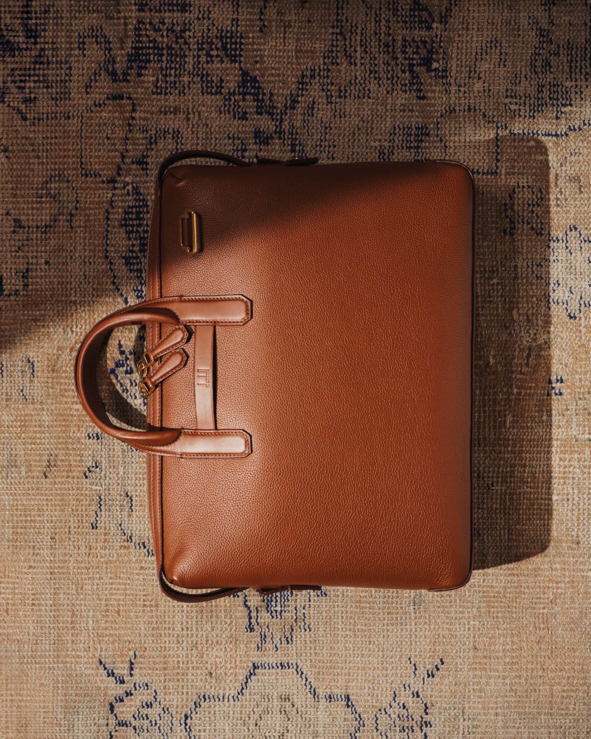 Overhead studio shot: a textured tan leather briefcase with a handle, resting on an ornate rug with blue and tan colors.