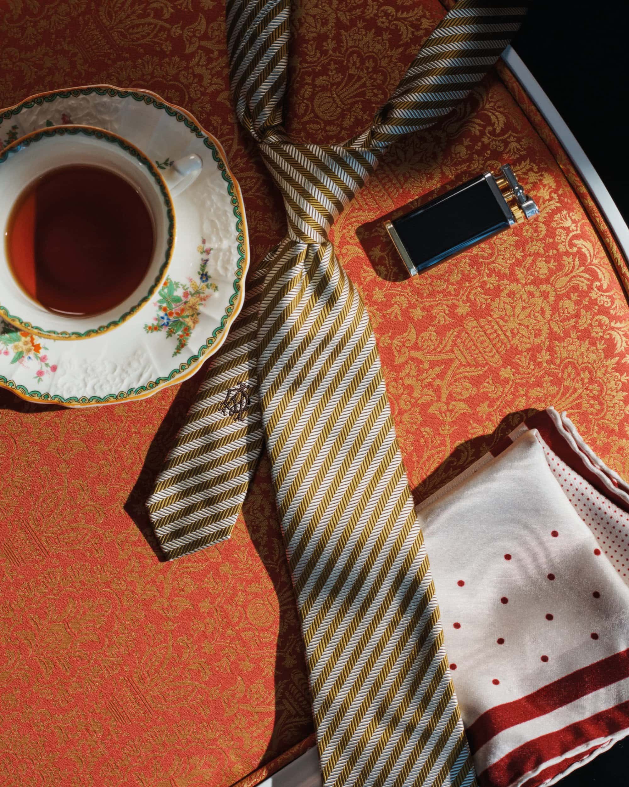 Still life shot of a striped tie, pocket square, teacup with saucer, and cigarette case on an ornate orange surface.