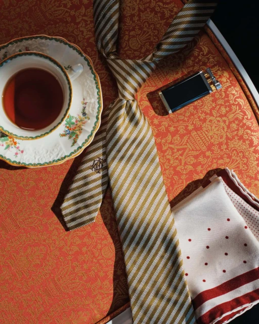 Still life shot of a striped tie, pocket square, teacup with saucer, and cigarette case on an ornate orange surface.