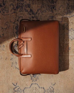 Brown leather briefcase resting flat on a patterned vintage textile rug with dramatic sunlight casting shadows.