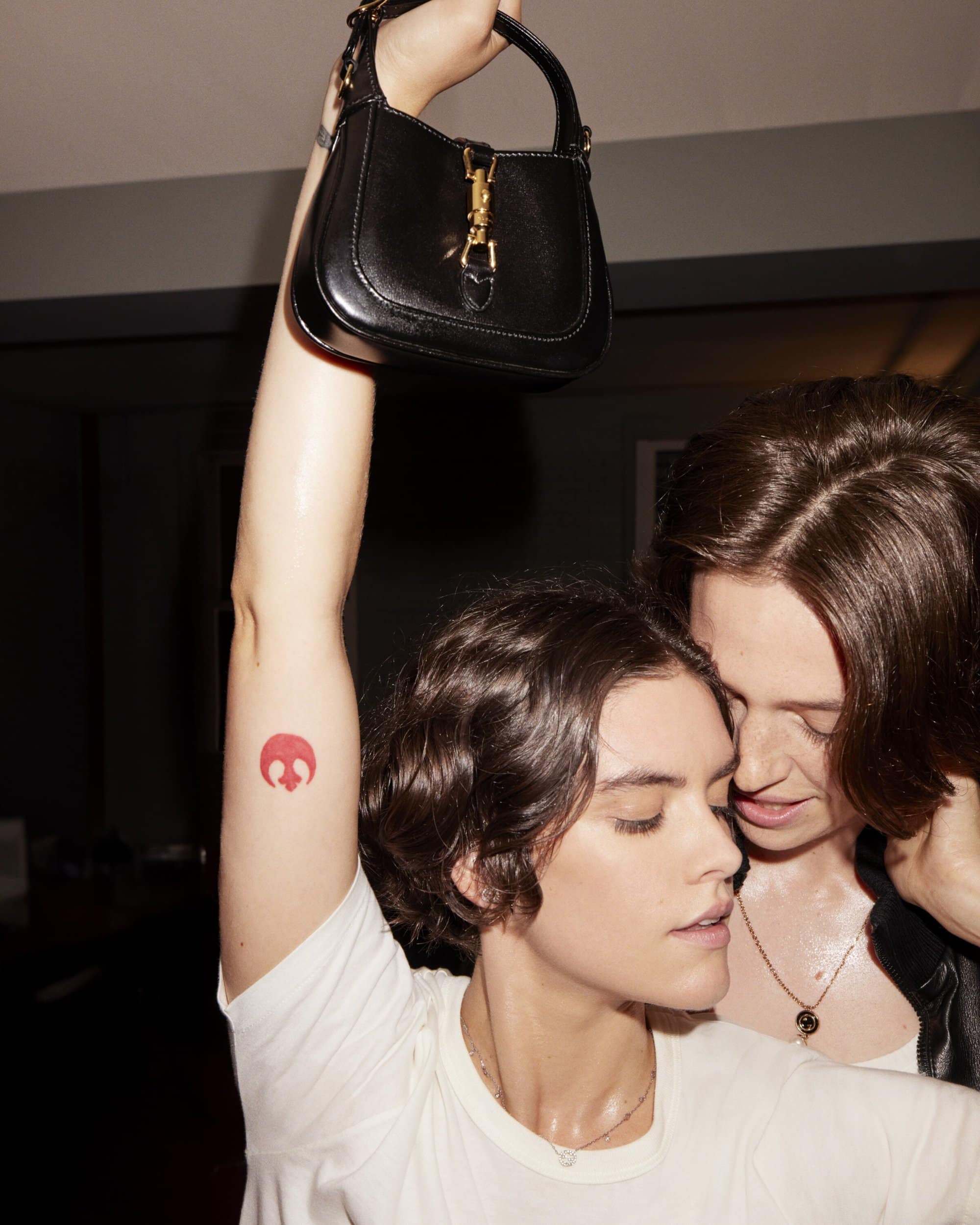 Two women pose indoors: one holding a black handbag above her head with a visible tattoo on her arm, and both wearing white t-shirts.