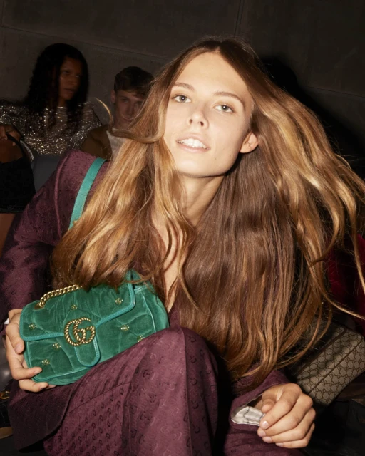 Blonde woman in a patterned suit holds a green velvet Gucci purse. Two people are visible in the background in a dim indoor setting.