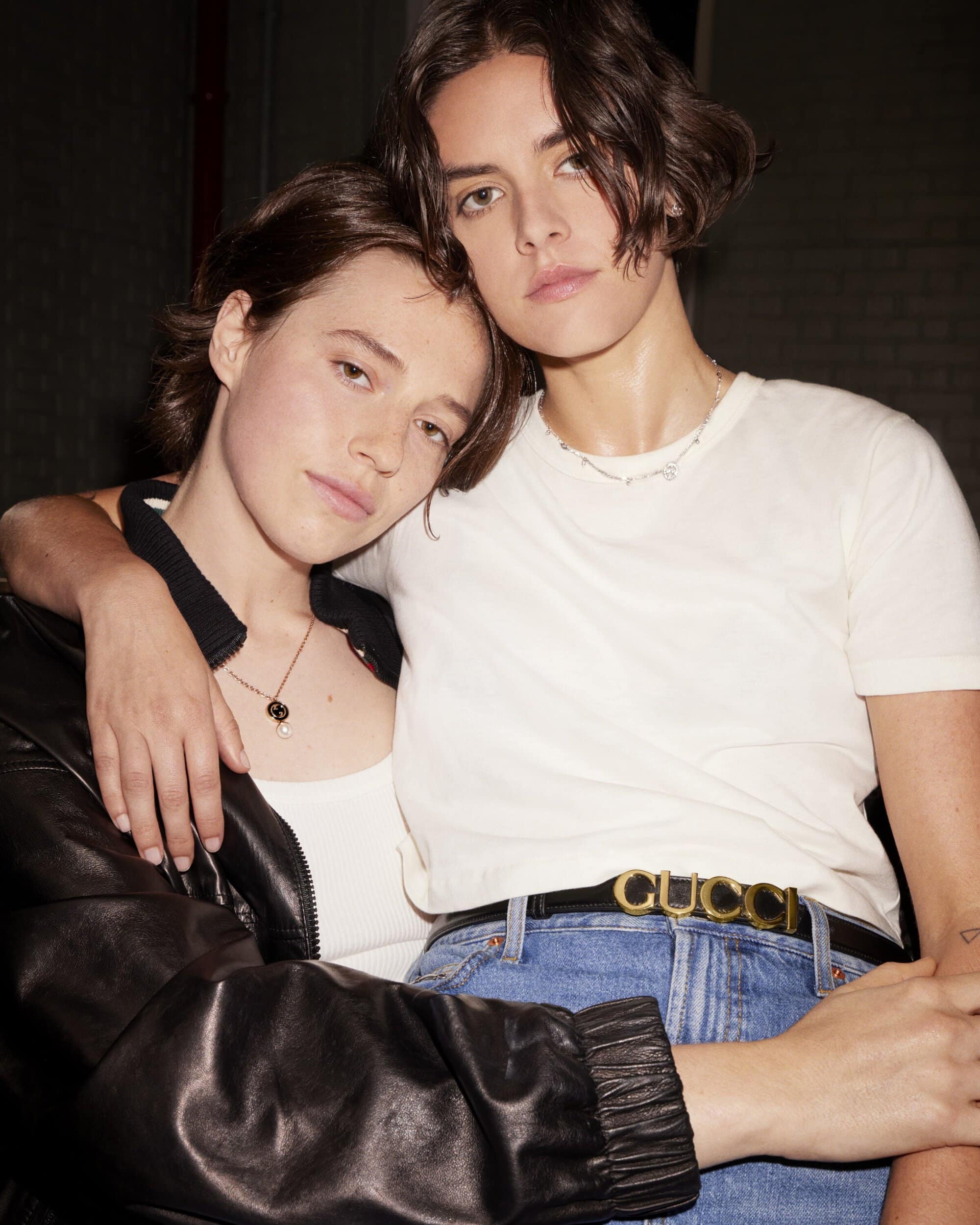 Two adult women embrace indoors, wearing casual clothing with a black leather jacket, t-shirt, jeans, and belts with logo details.