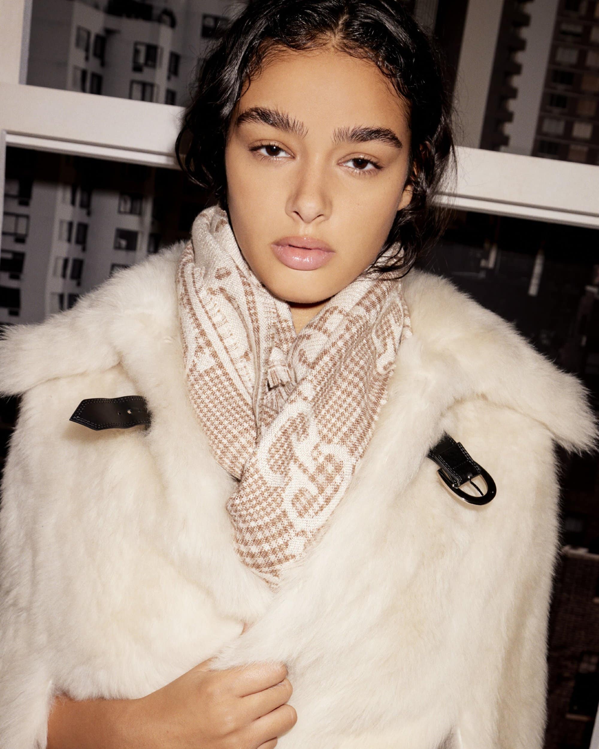 Model wearing a white fur coat and patterned scarf against an urban window backdrop