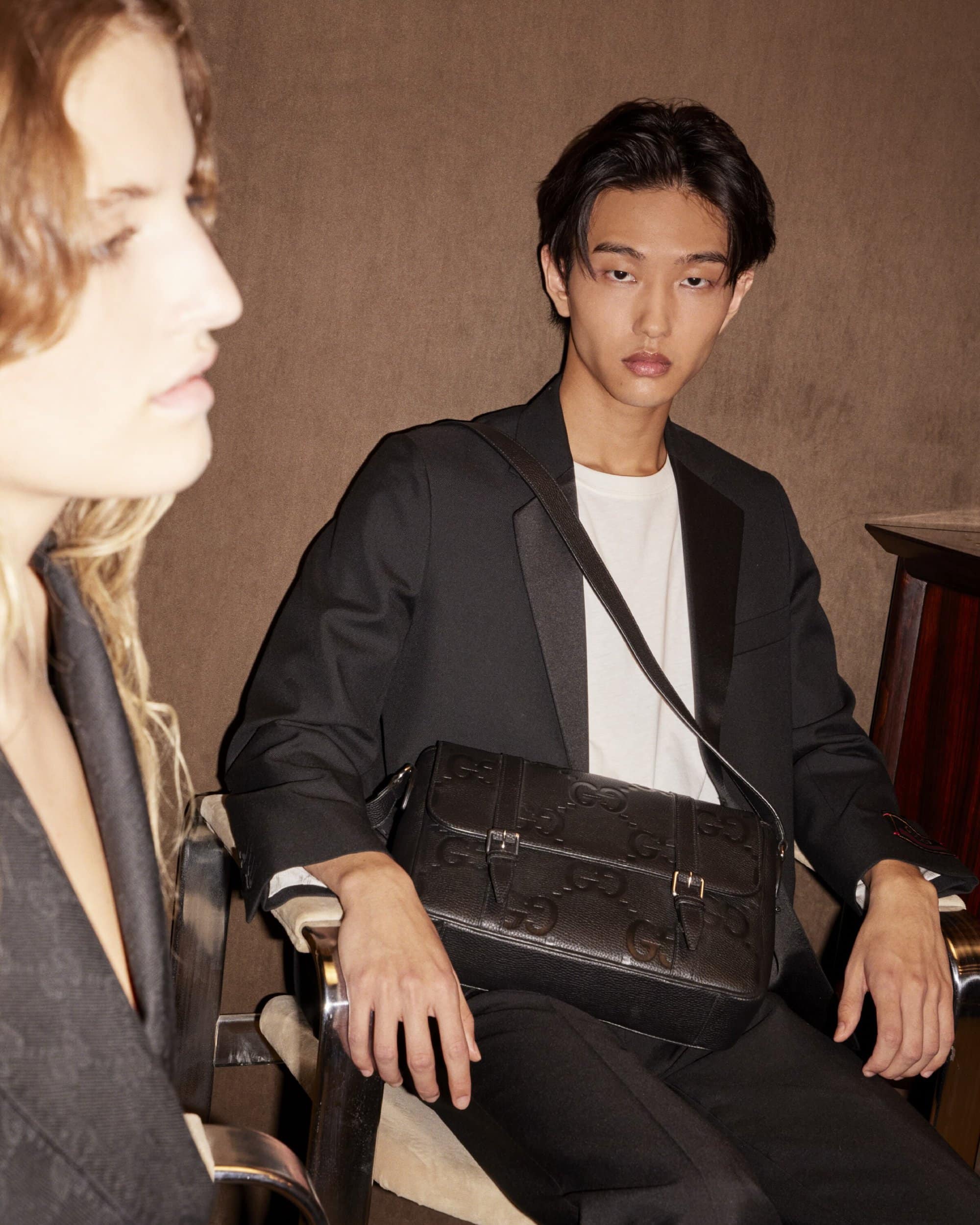 Fashionable man and woman modeling black blazers and a leather Gucci bag in an indoor setting with subdued lighting.