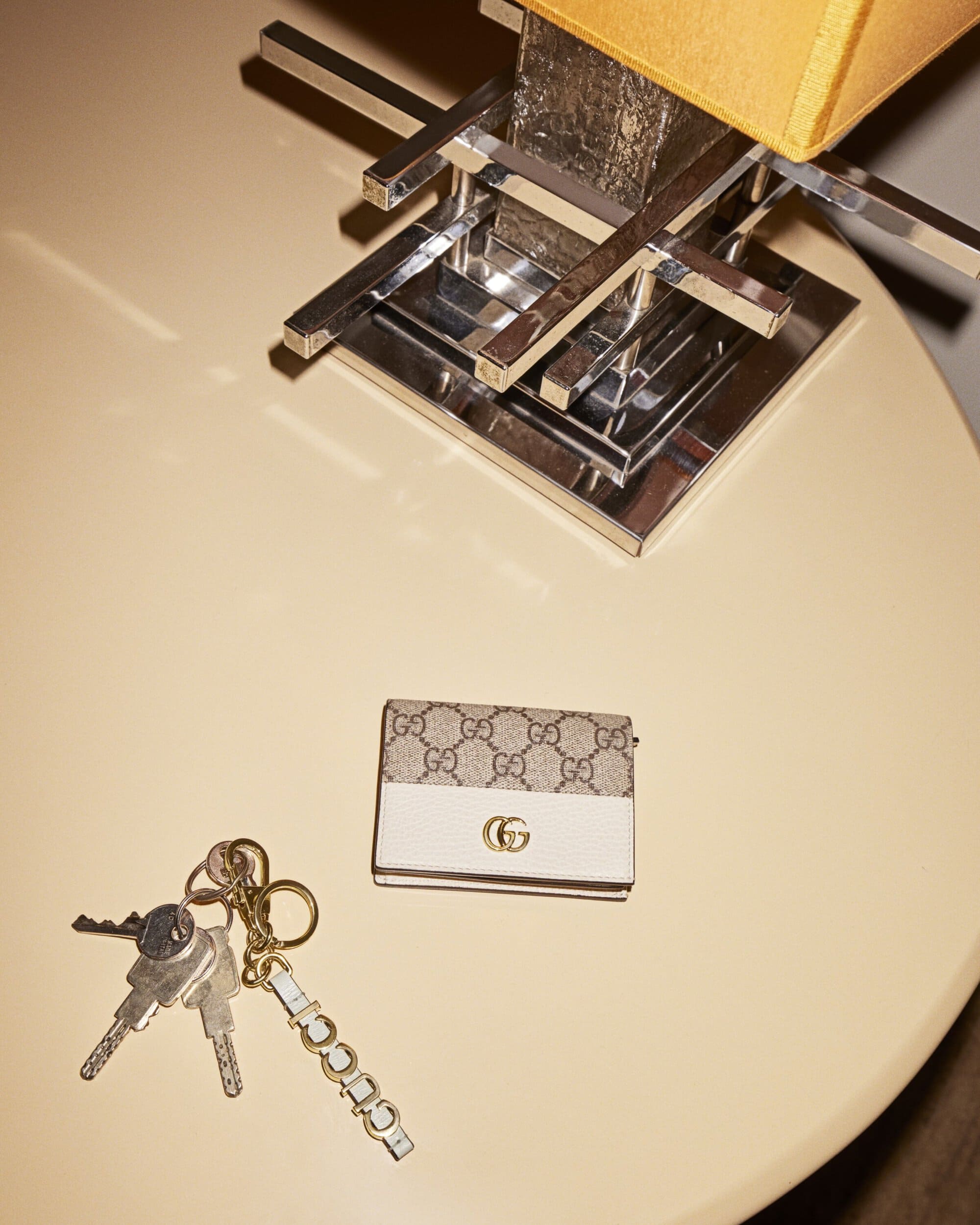 Overhead shot of a cream-colored table with a Gucci wallet, a set of keys, and a table lamp with a chrome base and mustard-colored shade.