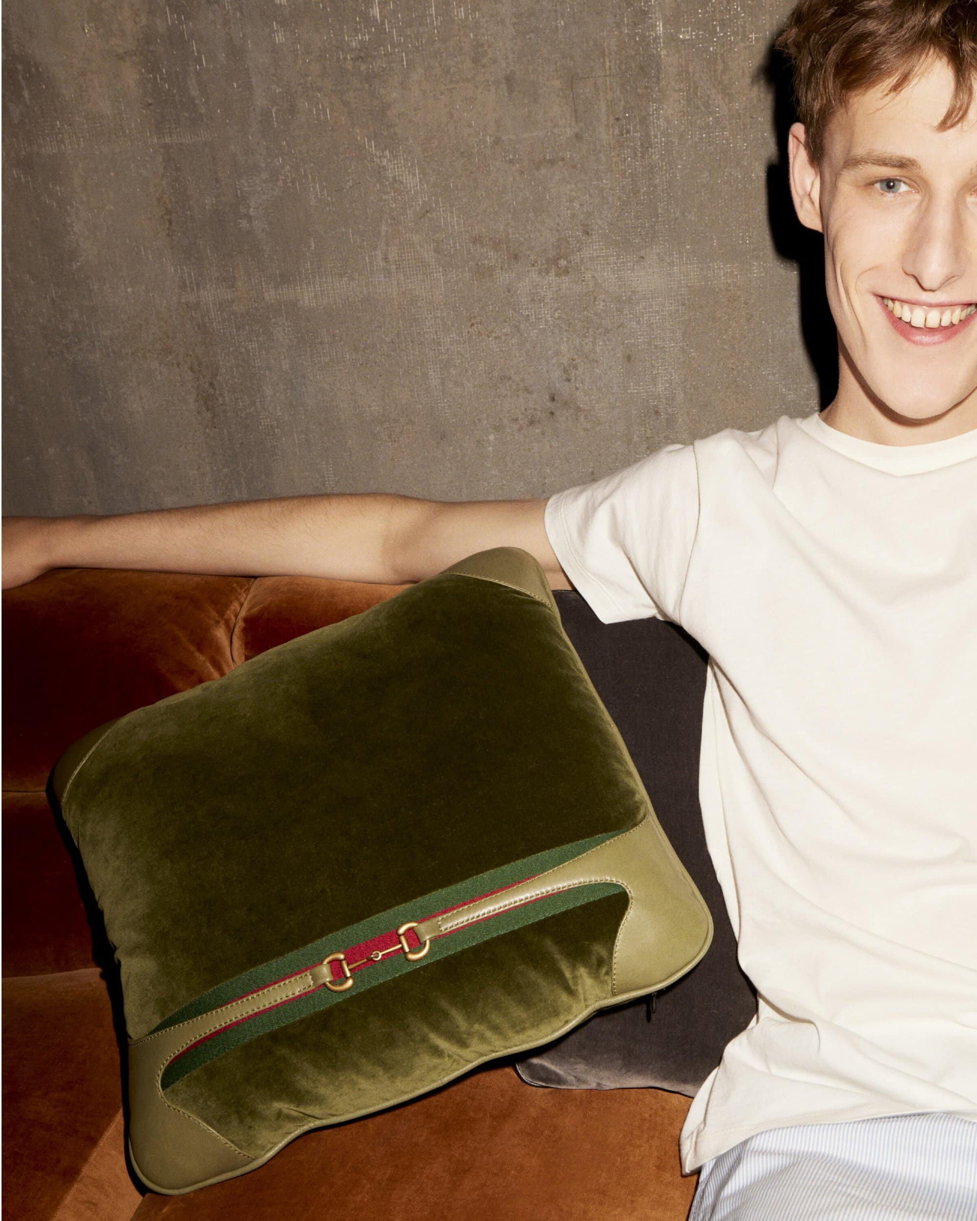 Smiling man on an orange couch with a green velvet pillow with a red and green band and metal horsebit detail, casual setting.