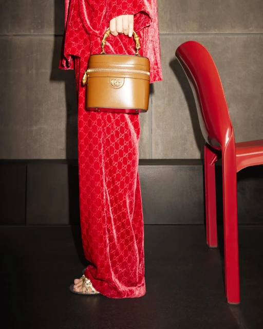 Woman in a red patterned suit holds a brown leather handbag with a bamboo handle next to a red chair and grey wall.