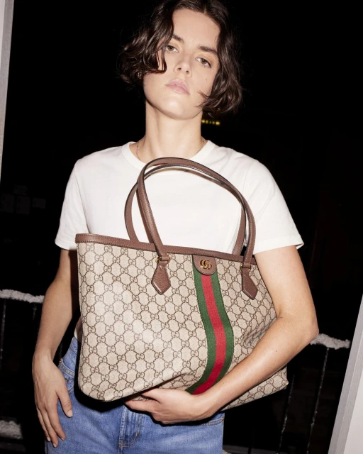 Adult female wearing a white shirt and blue jeans, holding a Gucci tote bag with red and green stripes in an indoor setting.